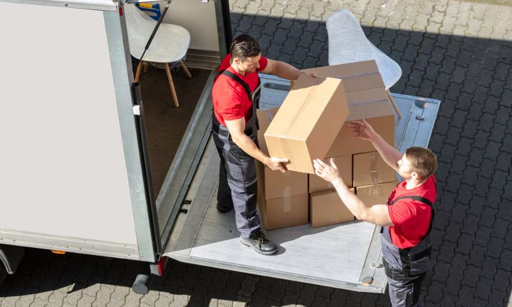 Two men, both of whom are wearing red shirts, are working together to take boxes out of the back of a moving truck.