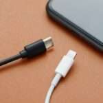 A close-up view shows a black USB-C charger and a white lightning charger sitting on a wooden surface by a smartphone.