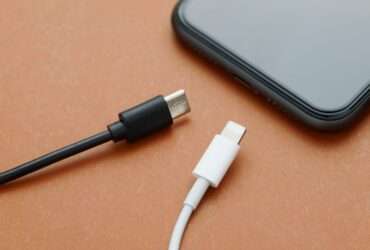 A close-up view shows a black USB-C charger and a white lightning charger sitting on a wooden surface by a smartphone.