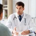 A doctor talking to a patient in their large office. The patient sits across a desk from the doctor.