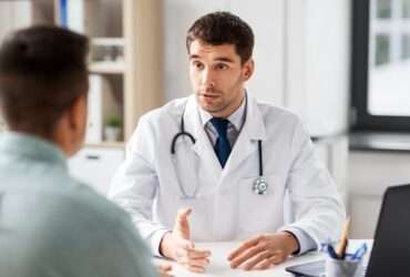 A doctor talking to a patient in their large office. The patient sits across a desk from the doctor.
