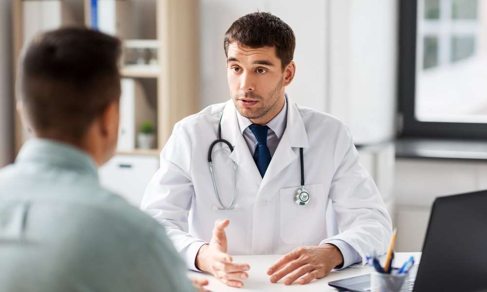 A doctor talking to a patient in their large office. The patient sits across a desk from the doctor.