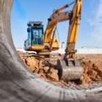A yellow excavator working behind a large metal pipe at a dusty construction site under a bright blue sky.