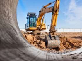 A yellow excavator working behind a large metal pipe at a dusty construction site under a bright blue sky.