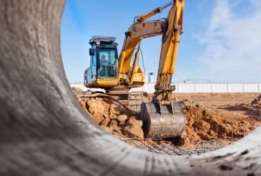 A yellow excavator working behind a large metal pipe at a dusty construction site under a bright blue sky.