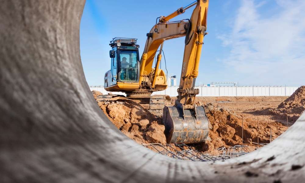 A yellow excavator working behind a large metal pipe at a dusty construction site under a bright blue sky.
