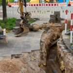 A yellow excavator digging a narrow trench beside a sidewalk, with safety barriers set up around the entire site.