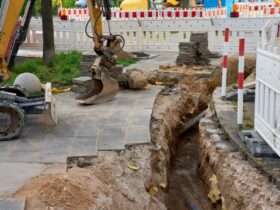 A yellow excavator digging a narrow trench beside a sidewalk, with safety barriers set up around the entire site.