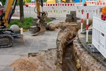 A yellow excavator digging a narrow trench beside a sidewalk, with safety barriers set up around the entire site.