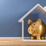 A white, house-shaped frame stands upright on plank flooring against a blue wall. A gold piggy bank is inside the house.