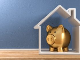 A white, house-shaped frame stands upright on plank flooring against a blue wall. A gold piggy bank is inside the house.