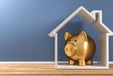A white, house-shaped frame stands upright on plank flooring against a blue wall. A gold piggy bank is inside the house.