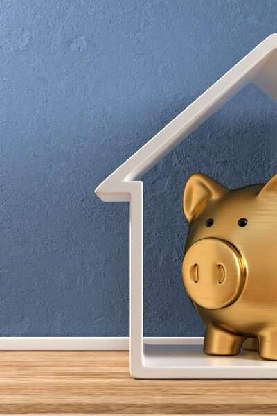 A white, house-shaped frame stands upright on plank flooring against a blue wall. A gold piggy bank is inside the house.