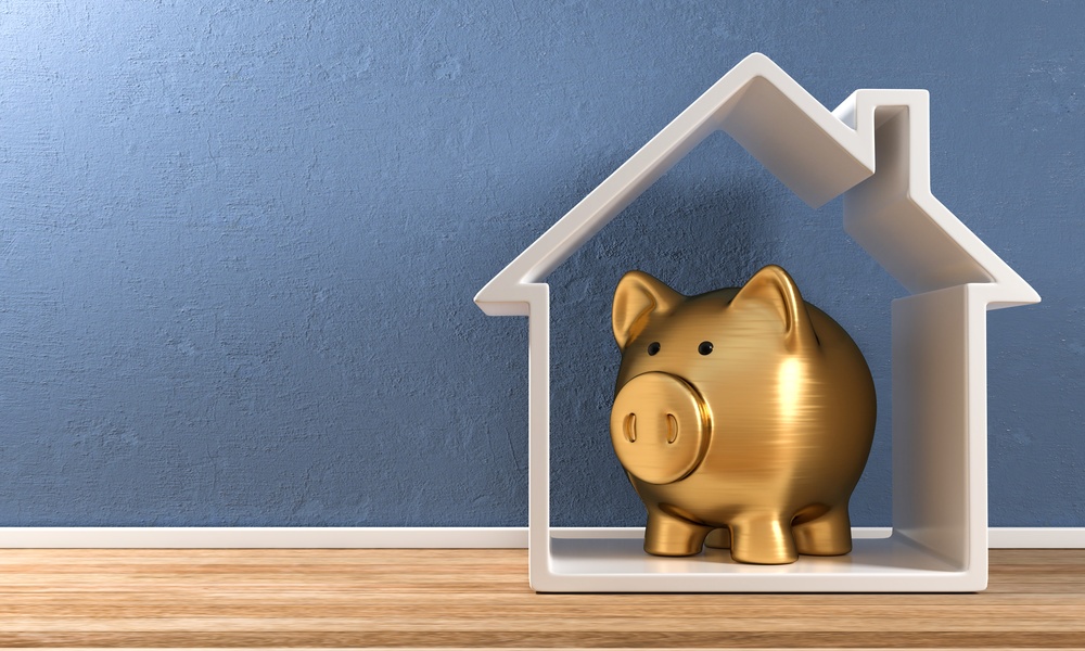 A white, house-shaped frame stands upright on plank flooring against a blue wall. A gold piggy bank is inside the house.