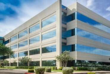 A large beige office building with blue reflective windows, trees, shrubs, and a cloudy sky beside a paved driveway.