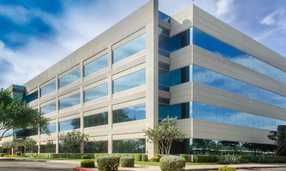 A large beige office building with blue reflective windows, trees, shrubs, and a cloudy sky beside a paved driveway.