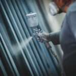 A worker wearing a respirator sprays protective coating onto metal pipes in an industrial workshop setting.