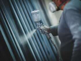 A worker wearing a respirator sprays protective coating onto metal pipes in an industrial workshop setting.