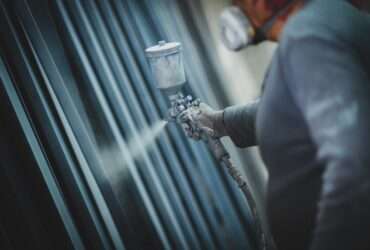 A worker wearing a respirator sprays protective coating onto metal pipes in an industrial workshop setting.