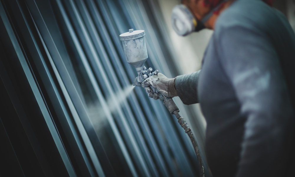 A worker wearing a respirator sprays protective coating onto metal pipes in an industrial workshop setting.