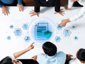 A group of people standing around a table with document and shield icons, pointing at a blue compliance graphic.