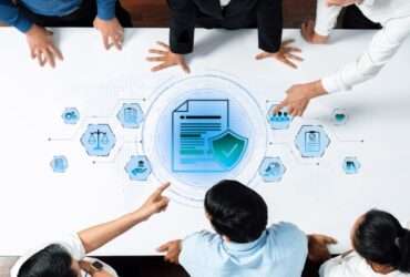 A group of people standing around a table with document and shield icons, pointing at a blue compliance graphic.