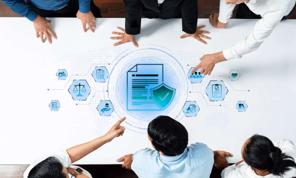 A group of people standing around a table with document and shield icons, pointing at a blue compliance graphic.