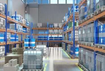A warehouse aisle lined with metal racks holding blue barrels, gray containers, and pallets on a polished floor.