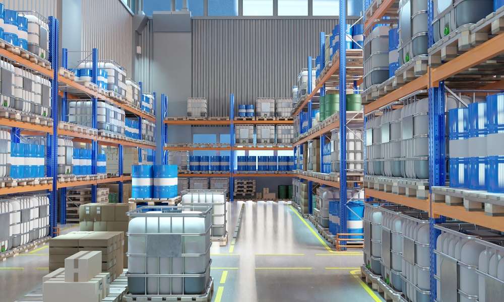 A warehouse aisle lined with metal racks holding blue barrels, gray containers, and pallets on a polished floor.