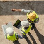 An aerial view shows three people wearing hard hats and yellow safety vest are looking at a laptop and construction plans.