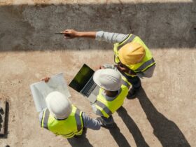 An aerial view shows three people wearing hard hats and yellow safety vest are looking at a laptop and construction plans.