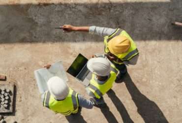 An aerial view shows three people wearing hard hats and yellow safety vest are looking at a laptop and construction plans.