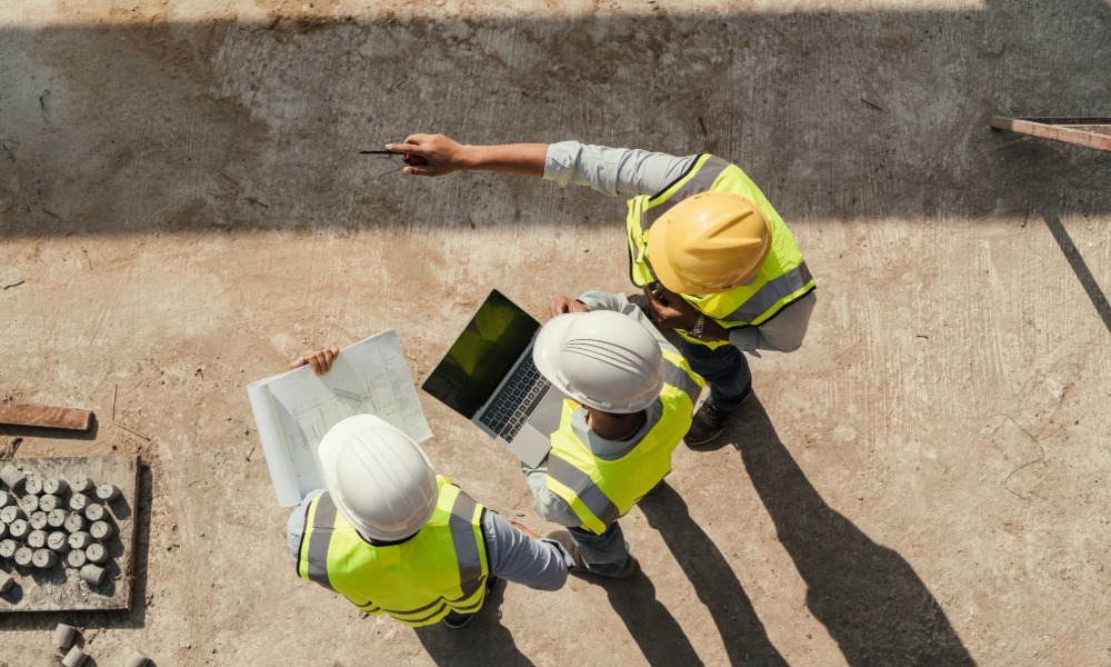 An aerial view shows three people wearing hard hats and yellow safety vest are looking at a laptop and construction plans.