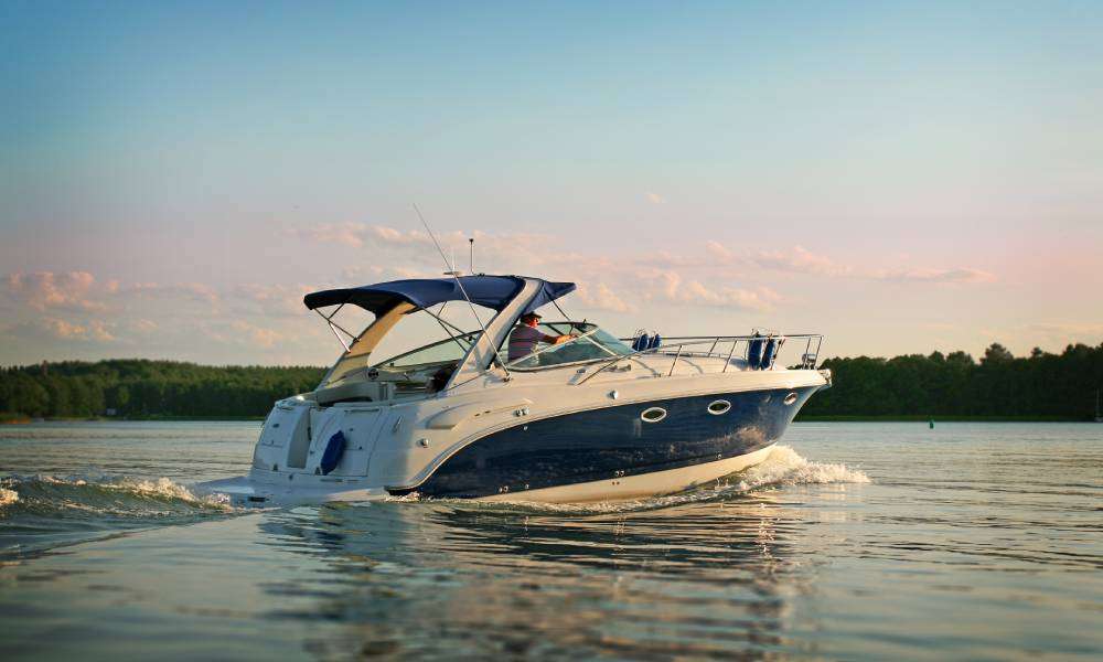 A man stands in his boat as it cruises through water. The sky has a few clouds, and a dense line of trees lines the shore.