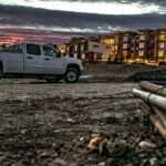 A truck is parked in the middle of a dirt road. There is an apartment building in the background.