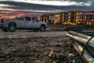 A truck is parked in the middle of a dirt road. There is an apartment building in the background.