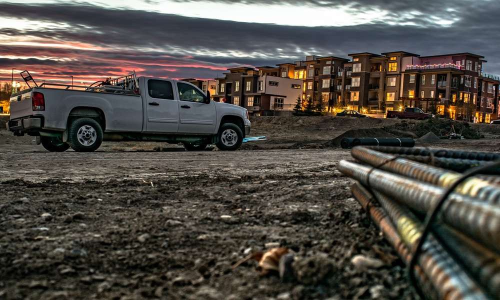 A truck is parked in the middle of a dirt road. There is an apartment building in the background.