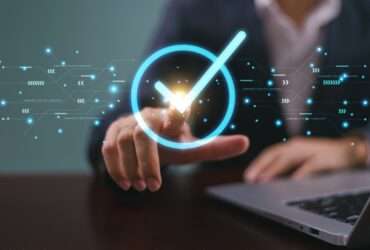 Close-up of a hand pressing a glowing check mark symbol with a laptop and digital interface graphics in view.