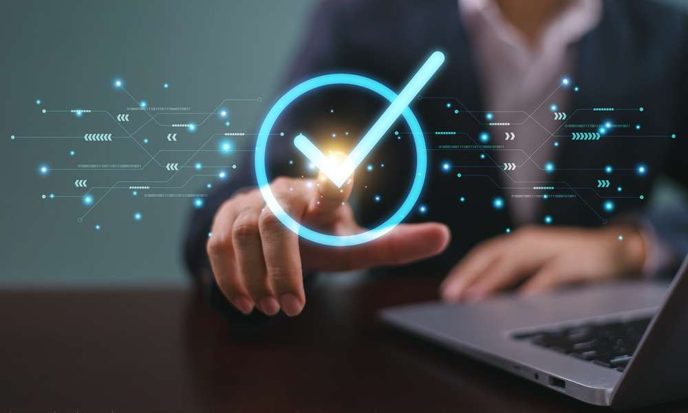 Close-up of a hand pressing a glowing check mark symbol with a laptop and digital interface graphics in view.