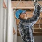 An electrician wearing a blue hard hat connects ceiling wiring inside a home under renovation with exposed wood beams.