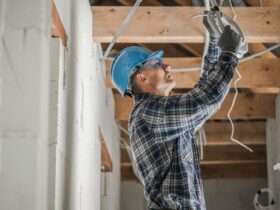 An electrician wearing a blue hard hat connects ceiling wiring inside a home under renovation with exposed wood beams.