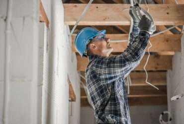 An electrician wearing a blue hard hat connects ceiling wiring inside a home under renovation with exposed wood beams.