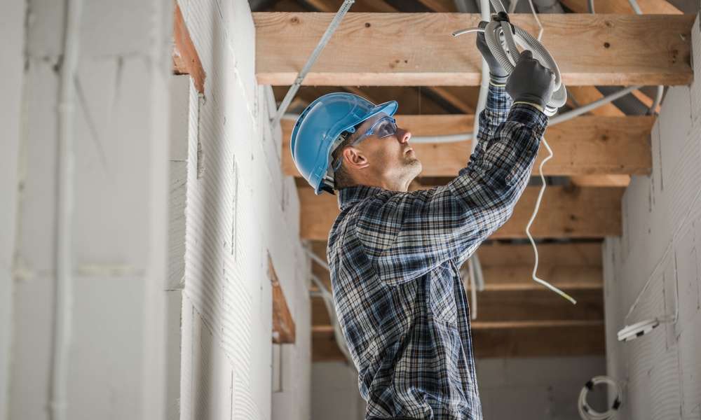 An electrician wearing a blue hard hat connects ceiling wiring inside a home under renovation with exposed wood beams.