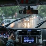 A woman driving a car on a rainy city street with windshield wipers, wet glass, headlights, and dashboard controls.