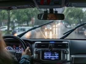A woman driving a car on a rainy city street with windshield wipers, wet glass, headlights, and dashboard controls.