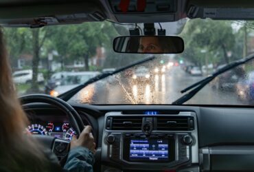 A woman driving a car on a rainy city street with windshield wipers, wet glass, headlights, and dashboard controls.