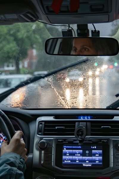 A woman driving a car on a rainy city street with windshield wipers, wet glass, headlights, and dashboard controls.