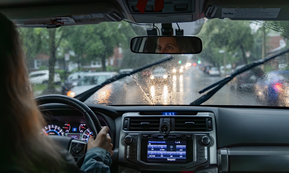 A woman driving a car on a rainy city street with windshield wipers, wet glass, headlights, and dashboard controls.