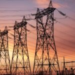 The sun is setting behind multiple high-voltage electrical transmission towers, with wires carrying electricity.