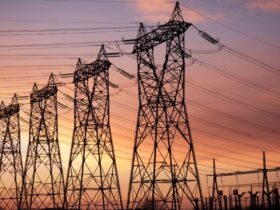 The sun is setting behind multiple high-voltage electrical transmission towers, with wires carrying electricity.